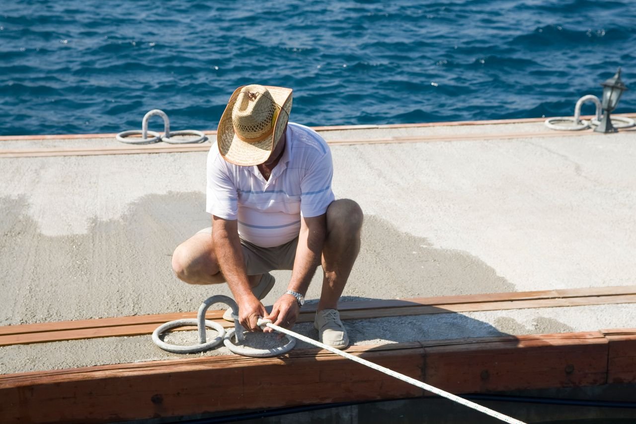 A man in a straw hat kneels on a dock, tying a boat rope under the bright sun.