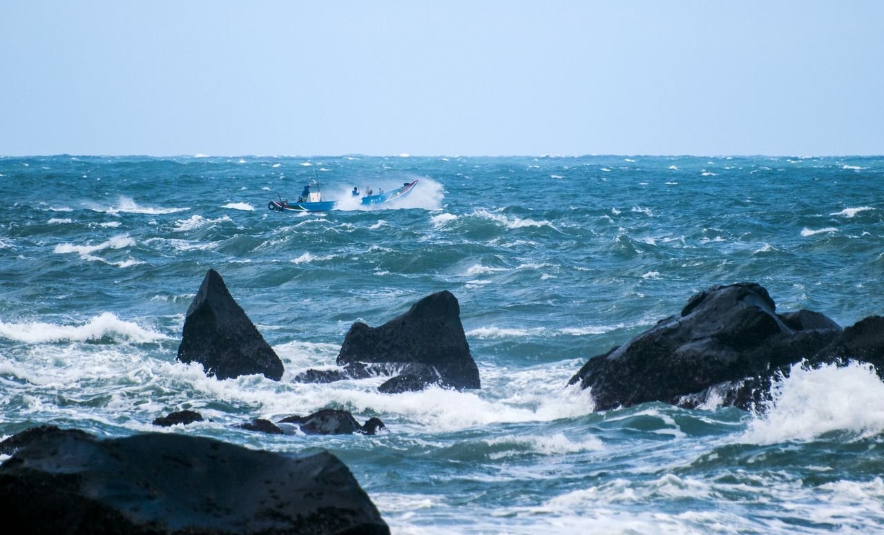 Formação A fishing boat navigates through rough seas near rocky shores under a bright day sky.